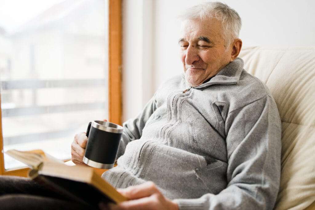 Senior man old sitting and Reading a book at the retirement nursing home with cup of tea in hand - The Preserve Senior male resident of the preserve at Dunedin enjoying a cup of coffee and reading a book